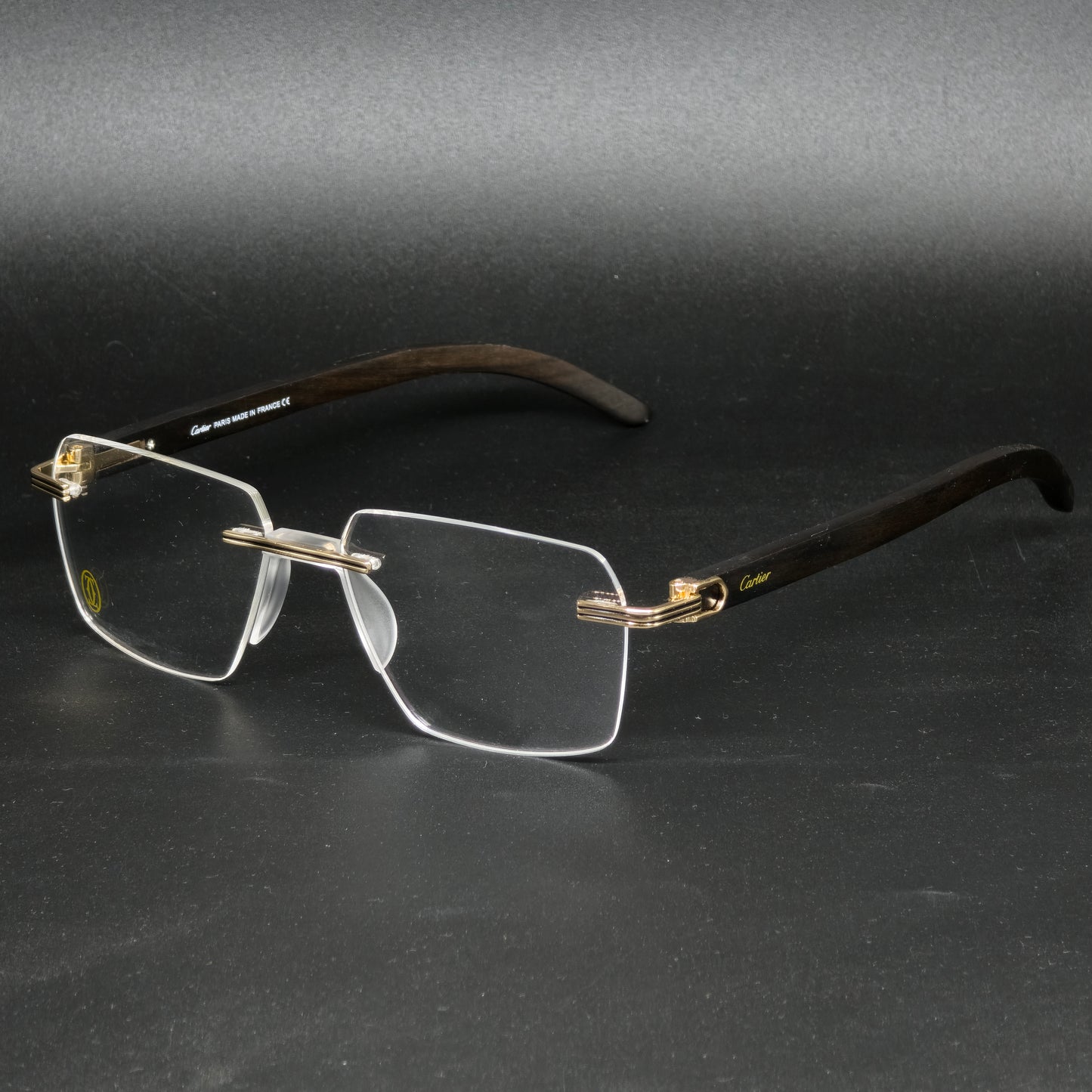 A pair of rimless eyeglasses with a dark brown wood-look frame, gold metal hinges, and clear lenses, displayed on a dark gray background.