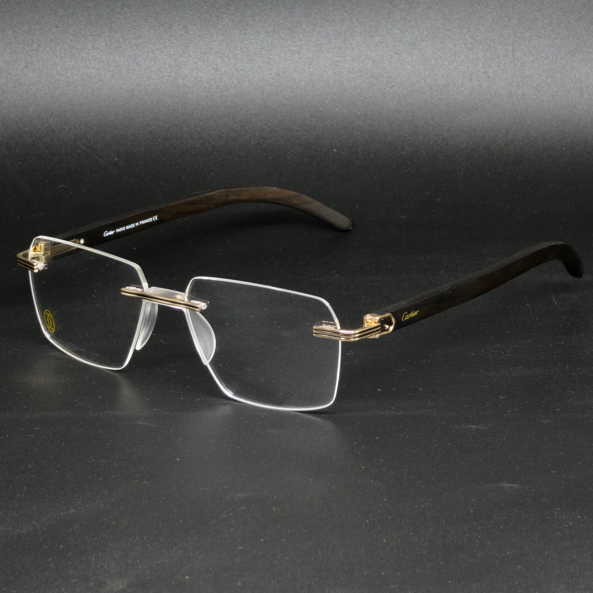 A pair of rimless eyeglasses with a dark brown wood-look frame, gold metal hinges, and clear lenses, displayed on a dark gray background.