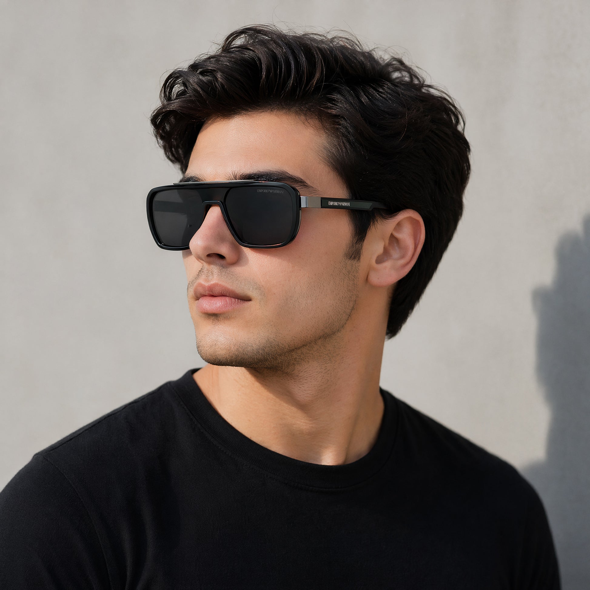 A man wearing black aviator-style sunglasses with a silver metal frame and dark lenses, photographed against a light gray textured wall.